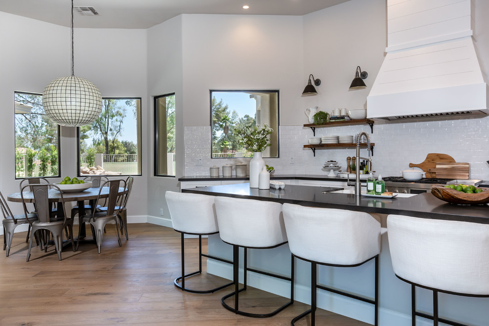 This is an example of a transitional l-shaped kitchen in Phoenix with shaker cabinets, white cabinets, white splashback, stainless steel appliances, medium hardwood floors, with island, brown floor and black benchtop.