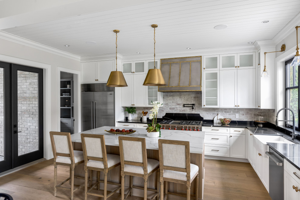 Large transitional l-shaped kitchen in Vancouver with a farmhouse sink, shaker cabinets, white cabinets, grey splashback, stainless steel appliances, medium hardwood floors, with island, beige floor and black benchtop.