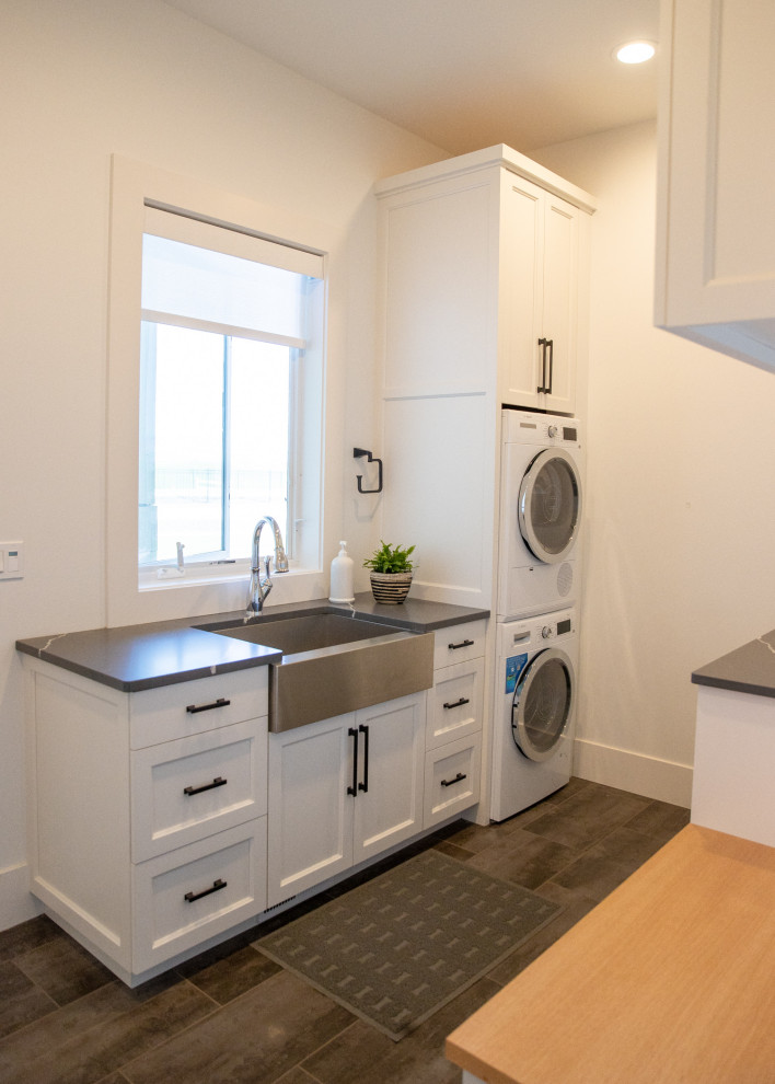 Laundry room - small modern ceramic tile and gray floor laundry room idea in Portland with a farmhouse sink, shaker cabinets, white cabinets, quartz countertops, white walls, a stacked washer/dryer and gray countertops