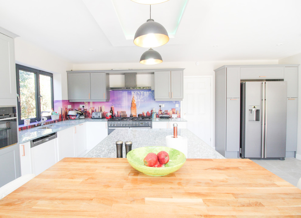 Expansive contemporary u-shaped open plan kitchen in London with an integrated sink, shaker cabinets, white cabinets, glass benchtops, multi-coloured splashback, glass sheet splashback, black appliances, ceramic floors, with island, grey floor and green benchtop.