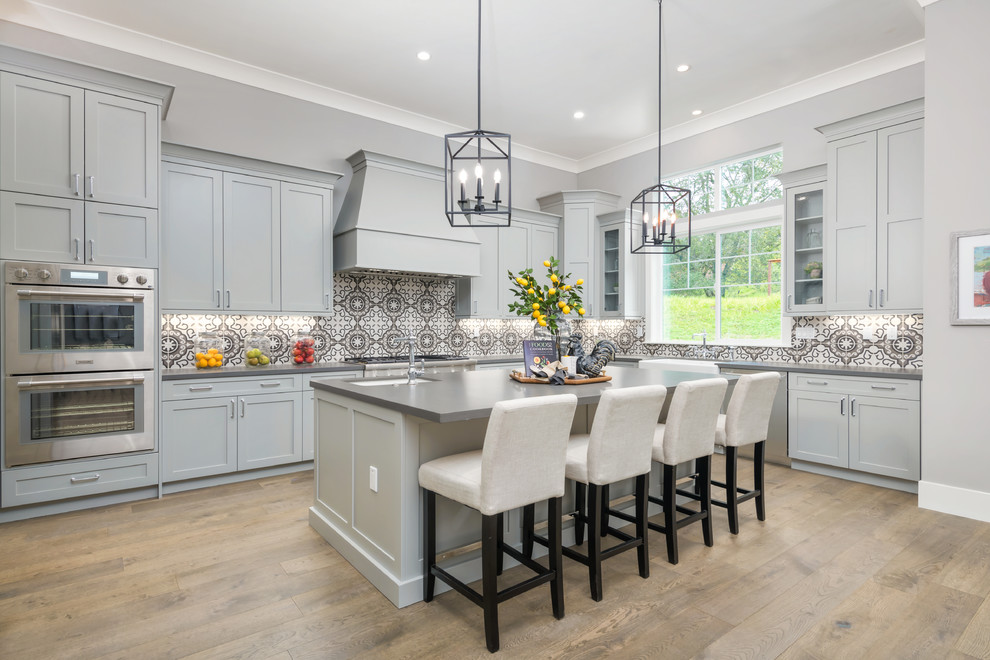 Example of a transitional l-shaped medium tone wood floor and brown floor kitchen design in San Francisco with shaker cabinets, gray cabinets, multicolored backsplash, stainless steel appliances, an island and gray countertops