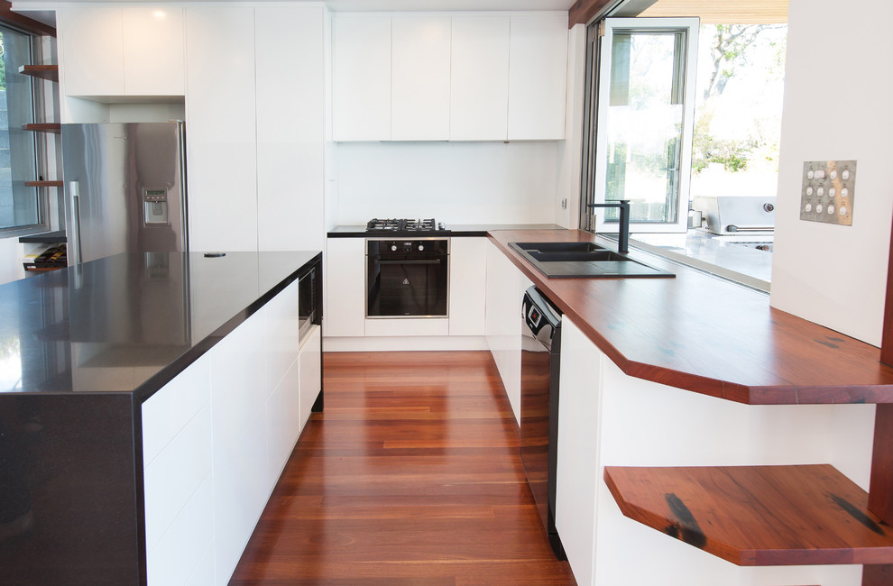 Design ideas for a mid-sized contemporary l-shaped open plan kitchen in Newcastle - Maitland with a drop-in sink, flat-panel cabinets, white cabinets, wood benchtops, white splashback, glass sheet splashback, black appliances, medium hardwood floors and with island.