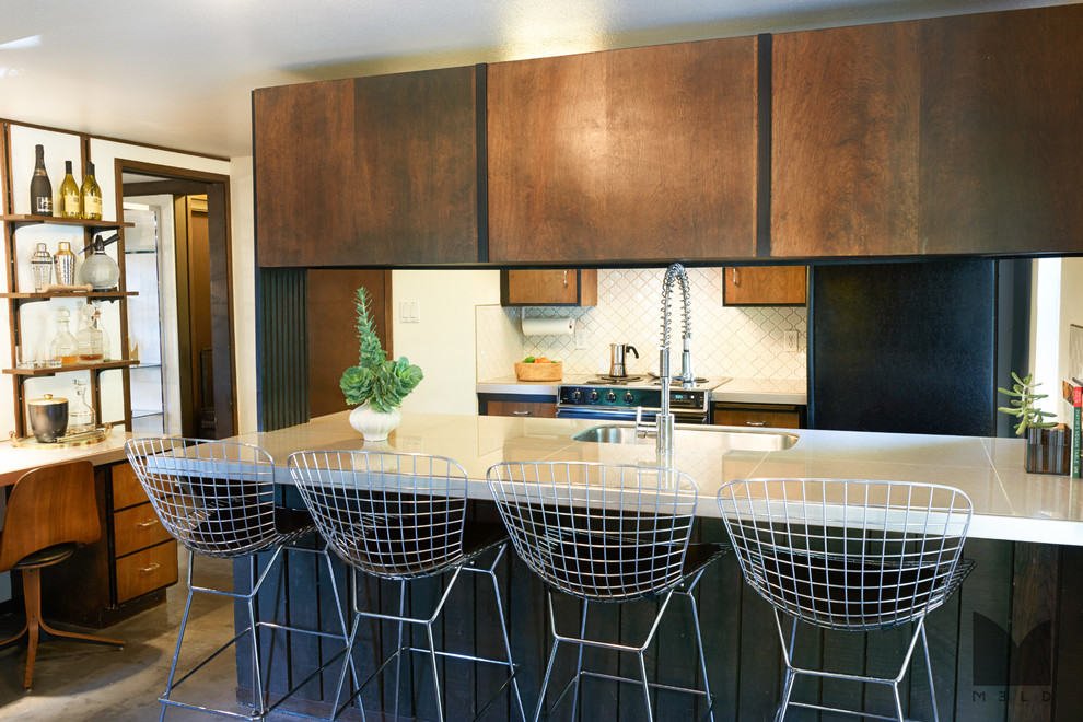 Small midcentury galley eat-in kitchen in Salt Lake City with a drop-in sink, flat-panel cabinets, medium wood cabinets, tile benchtops, white splashback, ceramic splashback, black appliances, concrete floors and a peninsula.