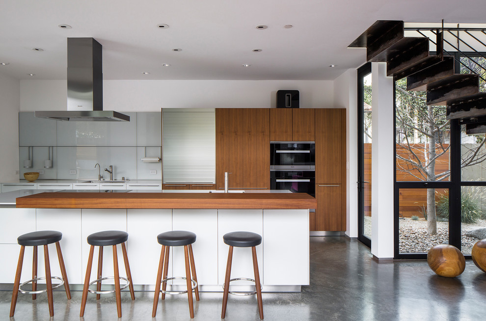 Inspiration for a modern galley eat-in kitchen in Los Angeles with a single-bowl sink, flat-panel cabinets, medium wood cabinets, grey splashback, glass sheet splashback, black appliances, concrete floors, with island and grey floor.