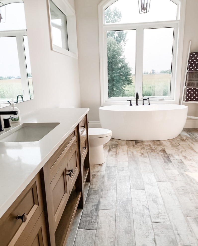 Inspiration for a large farmhouse ensuite bathroom in Chicago with light wood cabinets, a freestanding bath, a two-piece toilet, beige walls, wood-effect flooring, a submerged sink, engineered stone worktops, brown floors, beige worktops, a single sink, a freestanding vanity unit, a vaulted ceiling and recessed-panel cabinets.