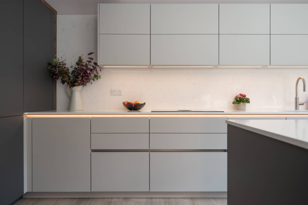 This is an example of a large contemporary l-shaped open plan kitchen in Hertfordshire with a double-bowl sink, flat-panel cabinets, white cabinets, quartzite benchtops, black appliances, with island, white benchtop and exposed beam.