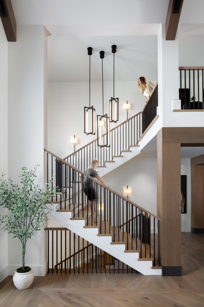 Luxury Custom Home Staircase Entryway Design - This grand staircase at the entry of this luxury custom home features light wood treads, black metal railings, and white risers for a mix of materiality and color contrast. Three industrial style pendants hang in the center at varying heights, and traditional style sconces line the walls. The staircase is finished with a large landing at the top for a reading nook.
