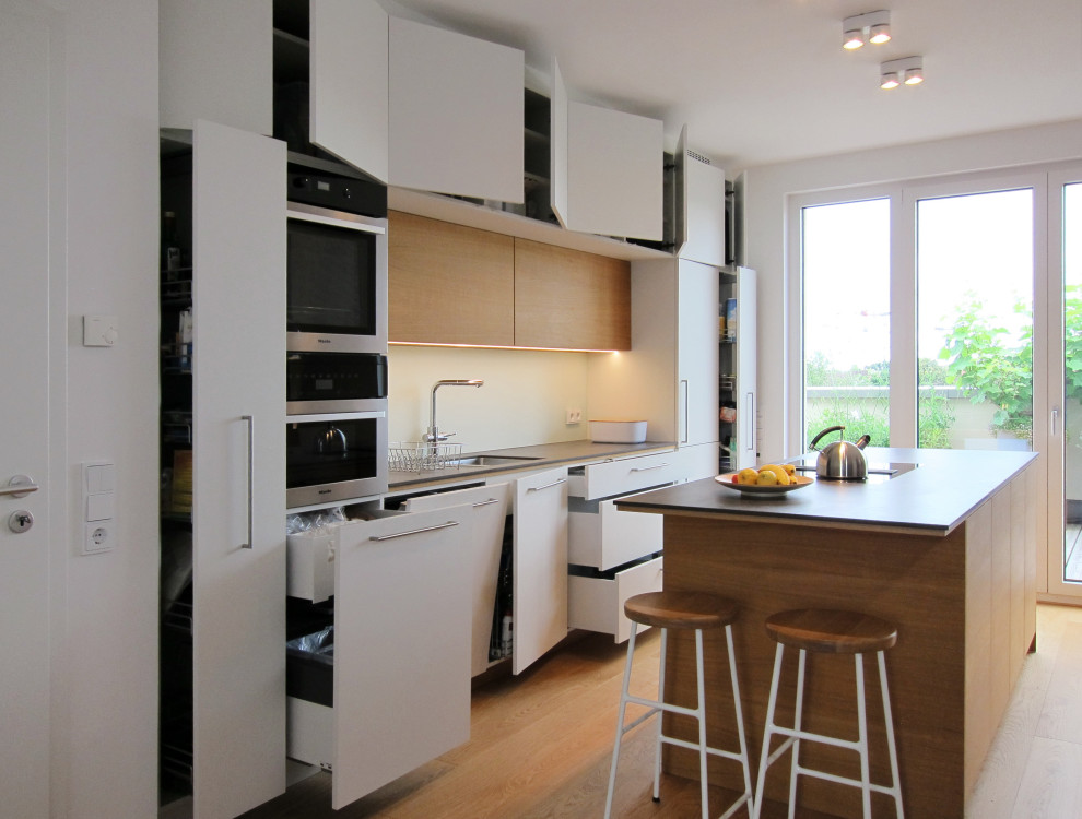 Inspiration for a large contemporary galley eat-in kitchen in Cologne with an undermount sink, flat-panel cabinets, white cabinets, tile benchtops, green splashback, glass sheet splashback, black appliances, light hardwood floors, with island and grey benchtop.