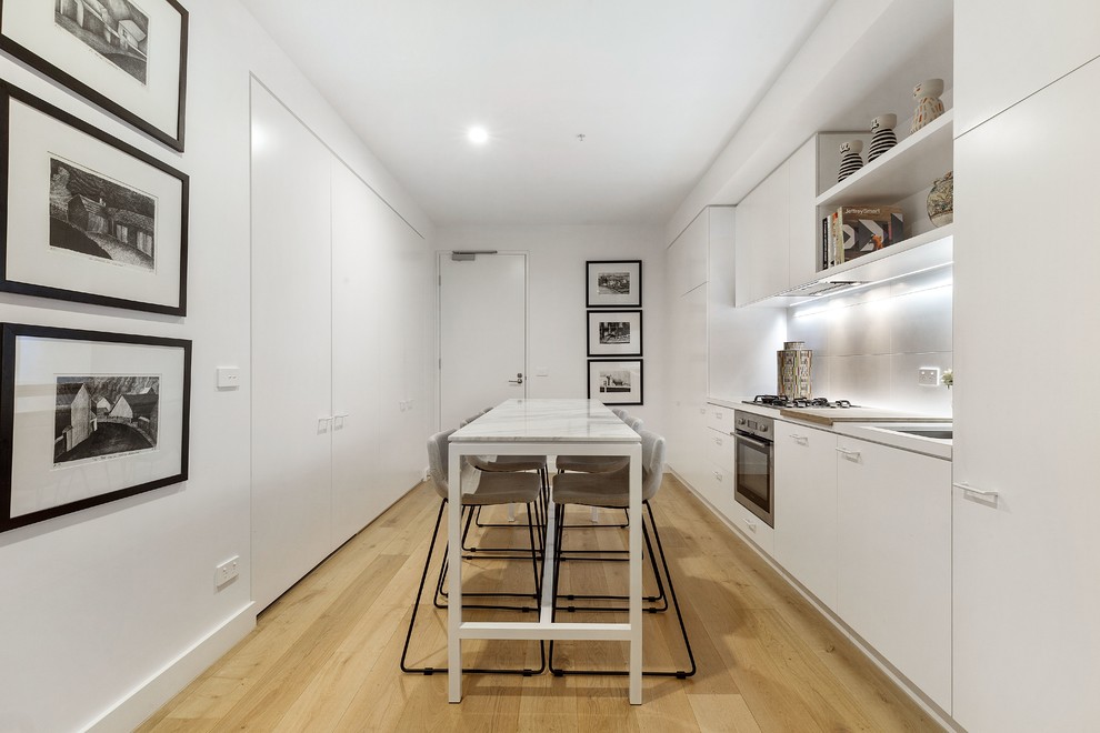 Photo of a contemporary single-wall kitchen in Melbourne with an undermount sink, flat-panel cabinets, white cabinets, stainless steel appliances, light hardwood floors, beige floor and white benchtop.