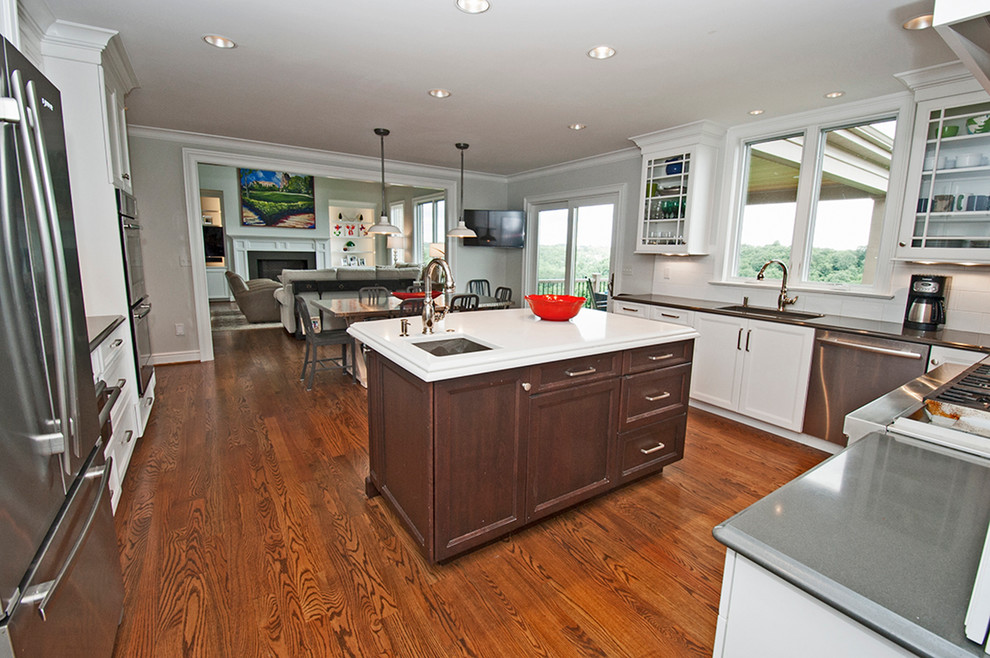 Example of a mid-sized transitional l-shaped medium tone wood floor open concept kitchen design with an undermount sink, glass-front cabinets, white cabinets, white backsplash, subway tile backsplash, stainless steel appliances and an island