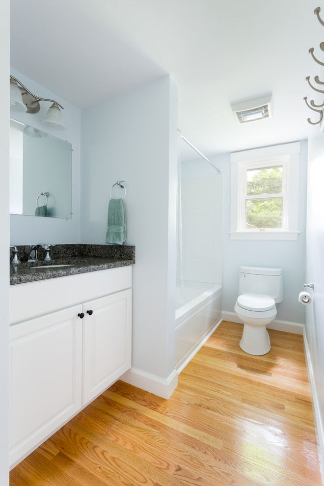Photo of a mid-sized traditional bathroom in Boston with raised-panel cabinets, white cabinets, an alcove tub, a shower/bathtub combo, a one-piece toilet, blue walls, light hardwood floors, an undermount sink, granite benchtops, a shower curtain and grey benchtops.