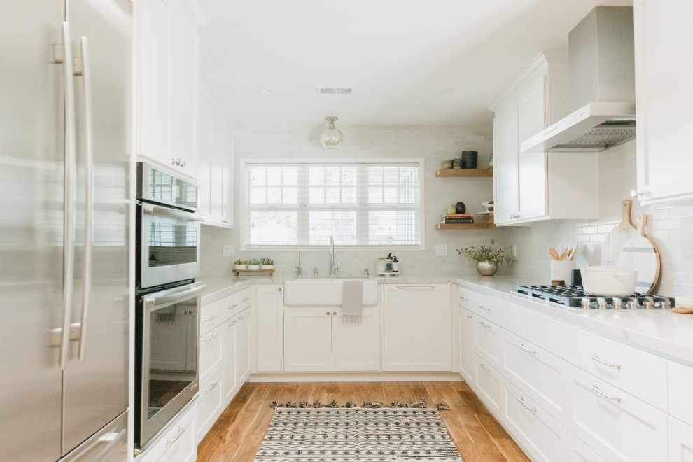 This is an example of a mid-sized transitional u-shaped eat-in kitchen in Sacramento with a farmhouse sink, shaker cabinets, white cabinets, quartzite benchtops, white splashback, ceramic splashback, stainless steel appliances, medium hardwood floors, no island, brown floor and white benchtop.