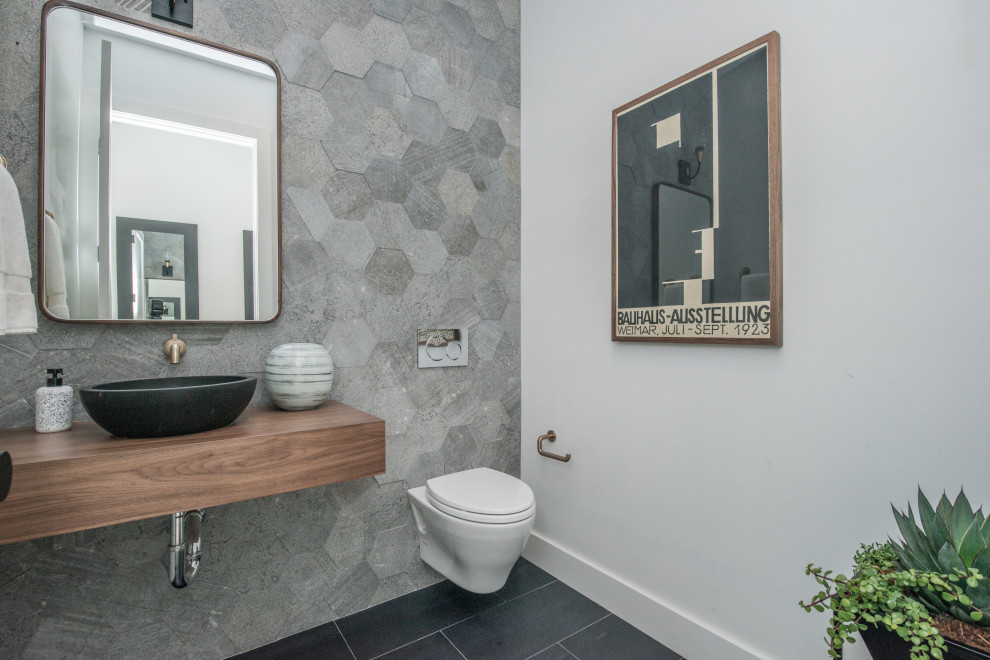 Small contemporary powder room in Los Angeles with open cabinets, medium wood cabinets, a wall-mount toilet, gray tile, cement tile, white walls, porcelain floors, a vessel sink, wood benchtops, black floor and a floating vanity.