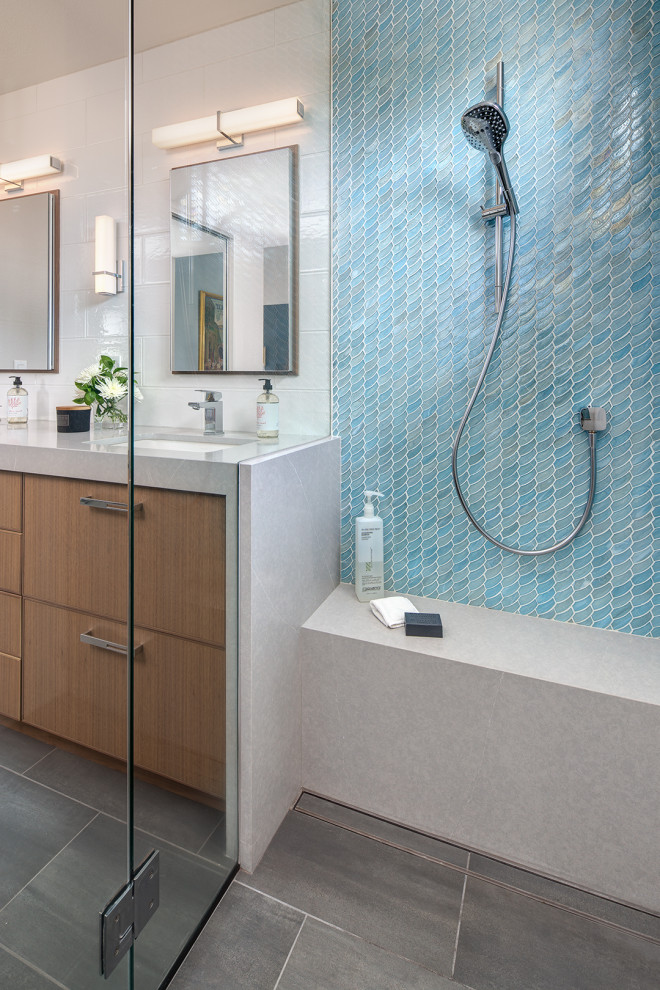 Cette photo montre une salle de bain principale bord de mer en bois clair de taille moyenne avec un placard à porte plane, une douche à l'italienne, un carrelage bleu, un carrelage en pâte de verre, un mur blanc, un sol en carrelage de porcelaine, un lavabo encastré, un plan de toilette en quartz modifié, un sol gris, une cabine de douche à porte battante, un plan de toilette gris, un banc de douche, meuble double vasque et meuble-lavabo encastré.