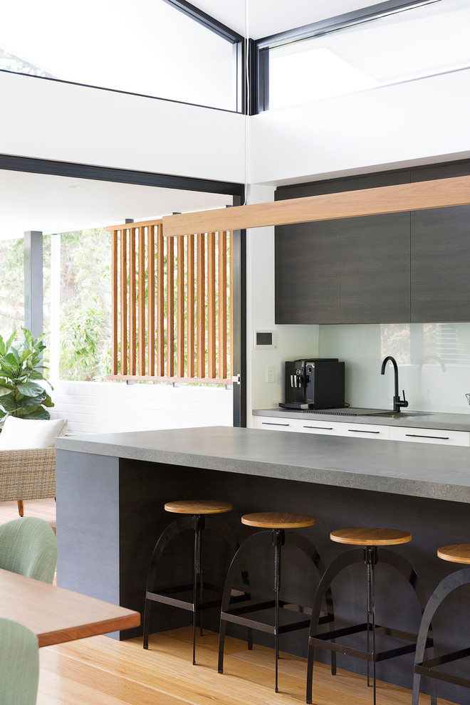 Design ideas for a mid-sized contemporary galley open plan kitchen in Sydney with an undermount sink, flat-panel cabinets, white cabinets, quartz benchtops, white splashback, glass sheet splashback, black appliances, medium hardwood floors, with island, brown floor and grey benchtop.