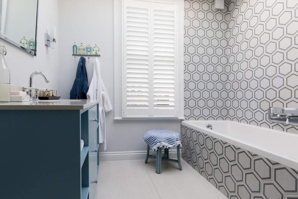 Photo of a mid-sized transitional kids bathroom in London with blue cabinets, a drop-in tub, gray tile, ceramic tile, grey walls, ceramic floors, engineered quartz benchtops, grey benchtops and grey floor.