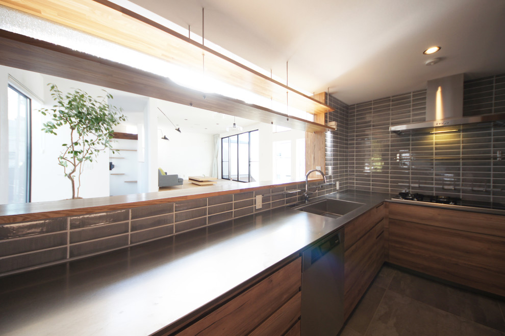 This is an example of a mid-sized contemporary l-shaped open plan kitchen in Tokyo with an integrated sink, flat-panel cabinets, medium wood cabinets, stainless steel benchtops, grey splashback, matchstick tile splashback, black appliances, slate floors and a peninsula.