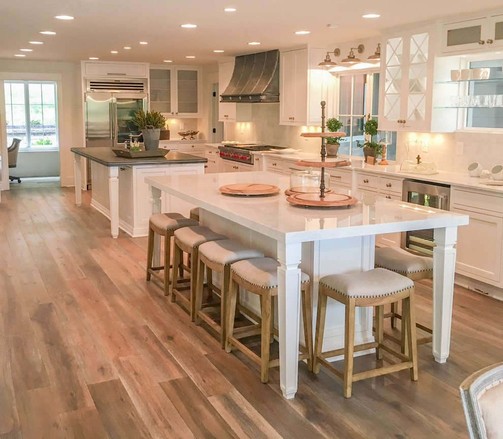 Design ideas for an expansive transitional l-shaped kitchen in Other with a farmhouse sink, beaded inset cabinets, white cabinets, zinc benchtops, white splashback, marble splashback, stainless steel appliances, light hardwood floors, multiple islands and brown floor.