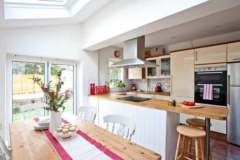This is an example of a transitional u-shaped eat-in kitchen in Surrey with a farmhouse sink, flat-panel cabinets, wood benchtops, white splashback, subway tile splashback, stainless steel appliances and a peninsula.