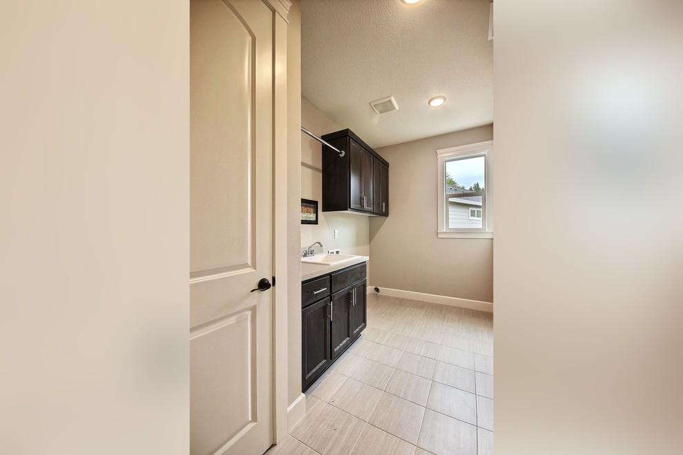 Photo of a mid-sized transitional single-wall utility room in Portland with a drop-in sink, recessed-panel cabinets, dark wood cabinets, tile benchtops, grey walls, travertine floors and a side-by-side washer and dryer.