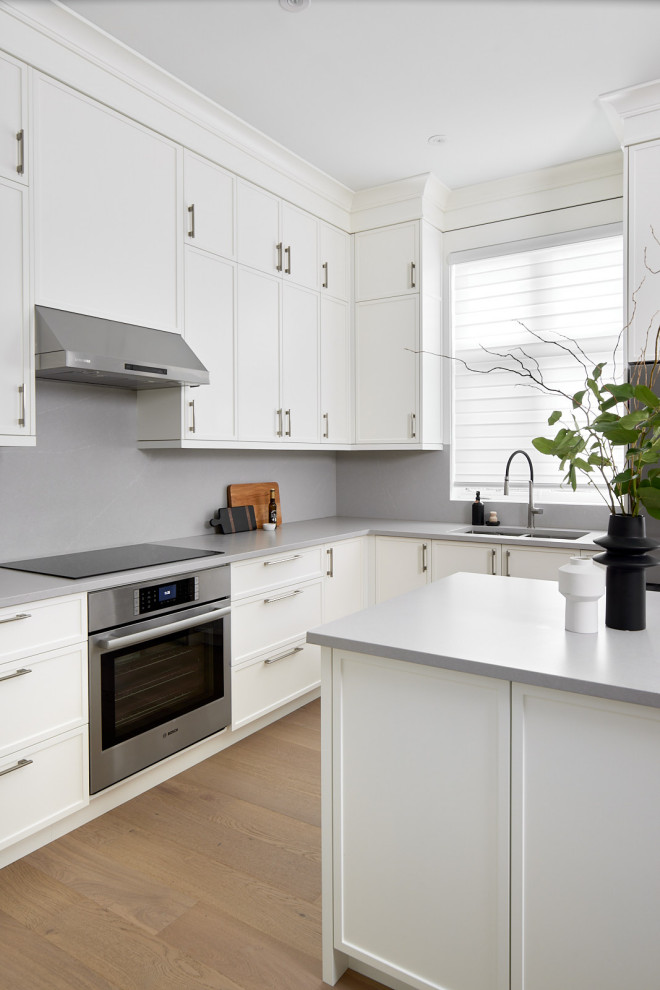 Example of a mid-sized transitional light wood floor kitchen design in Toronto with an undermount sink, shaker cabinets, white cabinets, quartz countertops, gray backsplash, quartz backsplash, stainless steel appliances and gray countertops