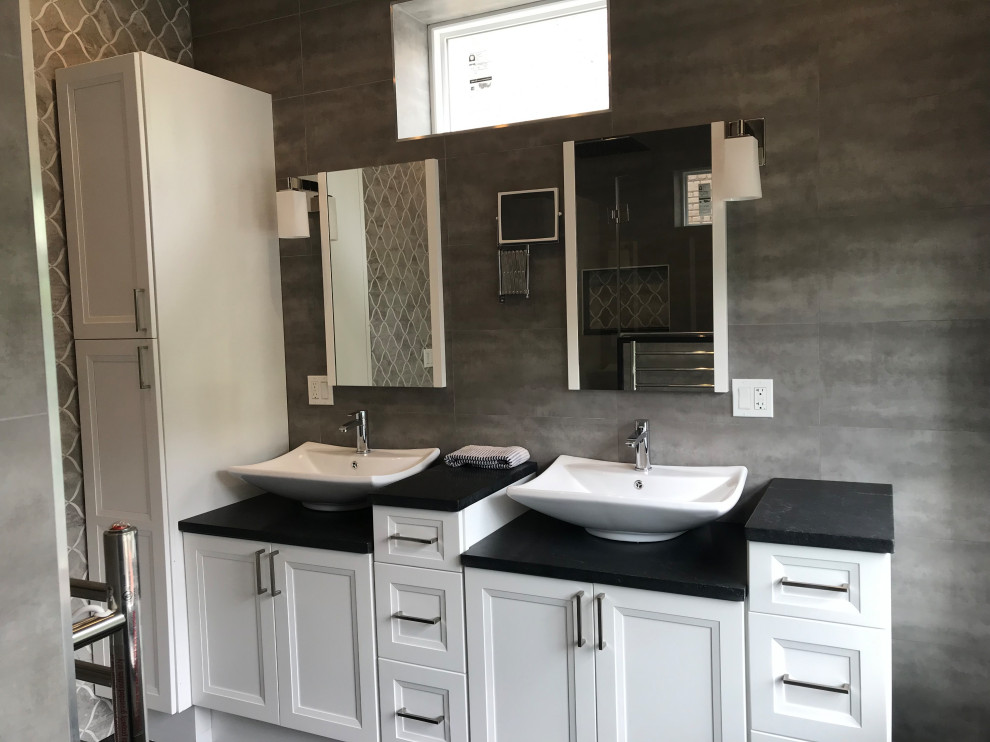 This is an example of a large contemporary master bathroom in New York with recessed-panel cabinets, white cabinets, gray tile, ceramic tile, granite benchtops, black benchtops, a double vanity, a built-in vanity, a corner tub, an alcove shower, a vessel sink, a hinged shower door, a two-piece toilet, green walls, vinyl floors, grey floor, a shower seat and vaulted.