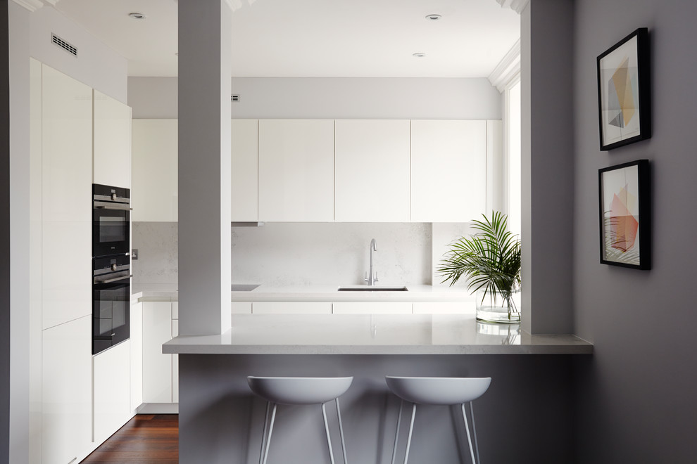 Design ideas for a small contemporary kitchen in London with an undermount sink, flat-panel cabinets, white cabinets, a peninsula, brown floor, grey splashback, black appliances and dark hardwood floors.