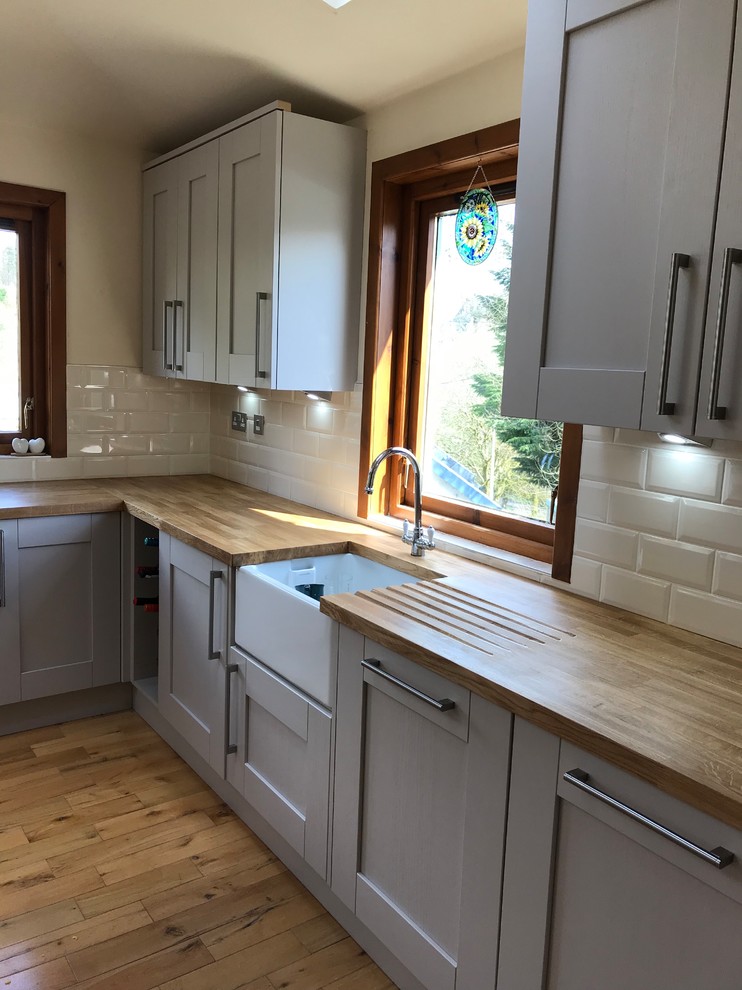 This is an example of a mid-sized traditional l-shaped eat-in kitchen in Glasgow with a farmhouse sink, shaker cabinets, wood benchtops, white splashback, subway tile splashback, black appliances, a peninsula, brown floor and brown benchtop.