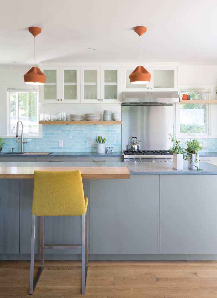 Contemporary l-shaped open plan kitchen in Austin with blue splashback, an undermount sink, flat-panel cabinets, grey cabinets, soapstone benchtops, matchstick tile splashback, stainless steel appliances, light hardwood floors, with island and grey benchtop.