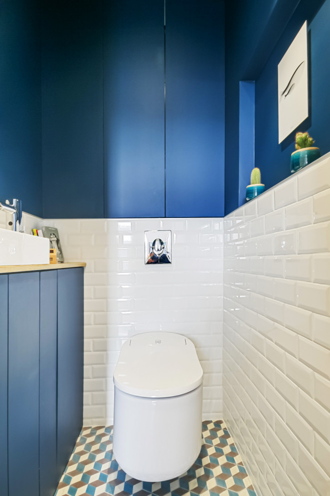 This is an example of a mid-sized contemporary powder room in Paris with beaded inset cabinets, a wall-mount toilet, white tile, subway tile, blue walls, cement tiles, a vessel sink, wood benchtops, blue floor, orange benchtops, white cabinets and a built-in vanity.