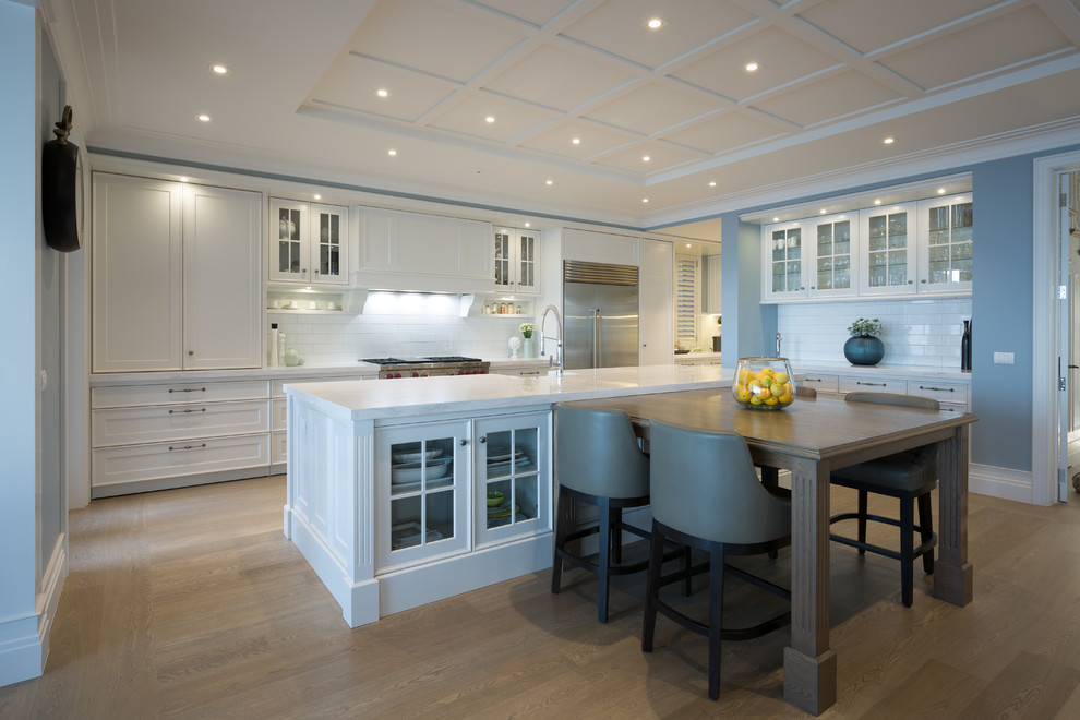 This is an example of a large beach style single-wall eat-in kitchen in Auckland with medium hardwood floors, a farmhouse sink, glass-front cabinets, white cabinets, white splashback, subway tile splashback, stainless steel appliances, with island and brown floor.