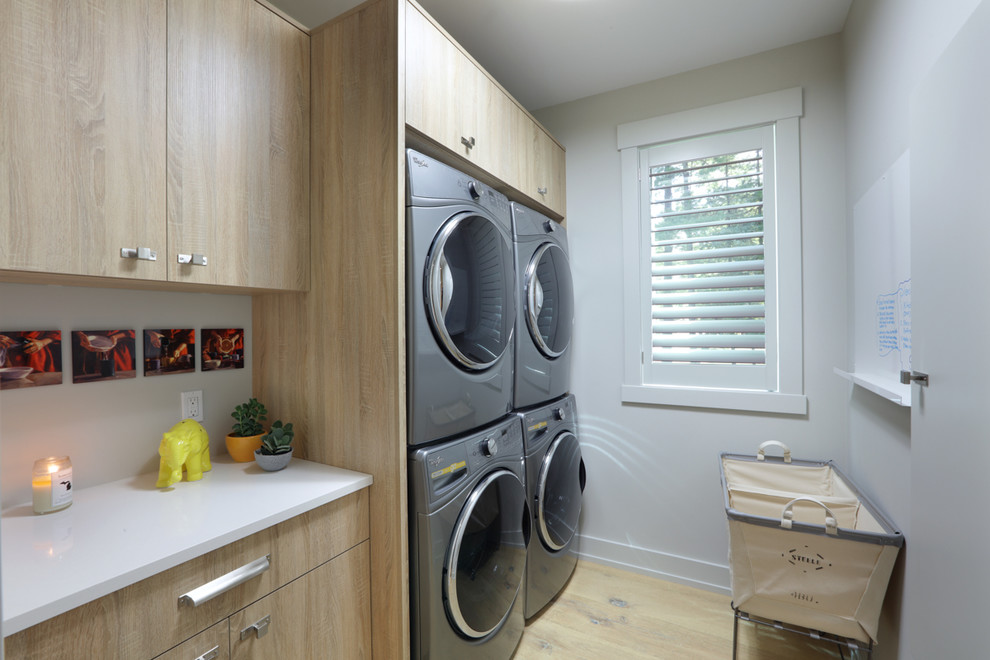 Inspiration for a mid-sized traditional single-wall dedicated laundry room in Grand Rapids with flat-panel cabinets, light wood cabinets, quartz benchtops, white walls, light hardwood floors, a stacked washer and dryer, brown floor and white benchtop.