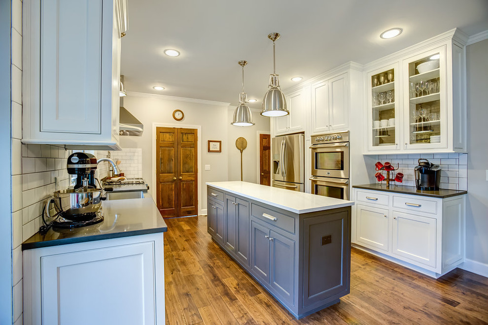Example of a mid-sized transitional galley medium tone wood floor eat-in kitchen design in Birmingham with a farmhouse sink, shaker cabinets, white cabinets, quartz countertops, white backsplash, subway tile backsplash, stainless steel appliances and an island