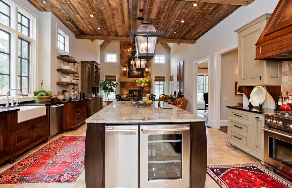 Example of a large mountain style l-shaped travertine floor kitchen design with a farmhouse sink, recessed-panel cabinets, beige cabinets, granite countertops, beige backsplash, stone tile backsplash, stainless steel appliances and an island