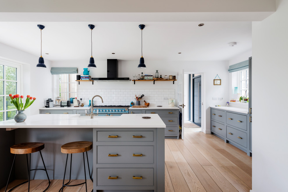 This is an example of a large transitional u-shaped open plan kitchen in Devon with a farmhouse sink, beaded inset cabinets, grey cabinets, solid surface benchtops, white splashback, porcelain splashback, coloured appliances, medium hardwood floors, with island, white benchtop and beige floor.
