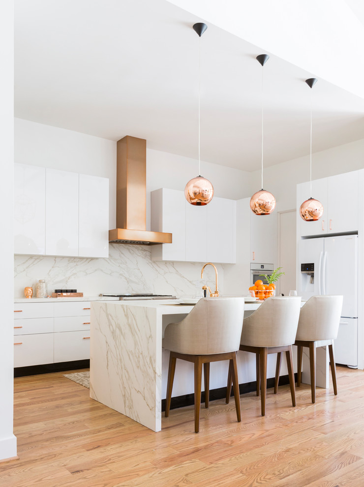 Photo of a contemporary l-shaped kitchen in Houston with flat-panel cabinets, white cabinets, white splashback, stone slab splashback, white appliances, medium hardwood floors, with island, brown floor and white benchtop.