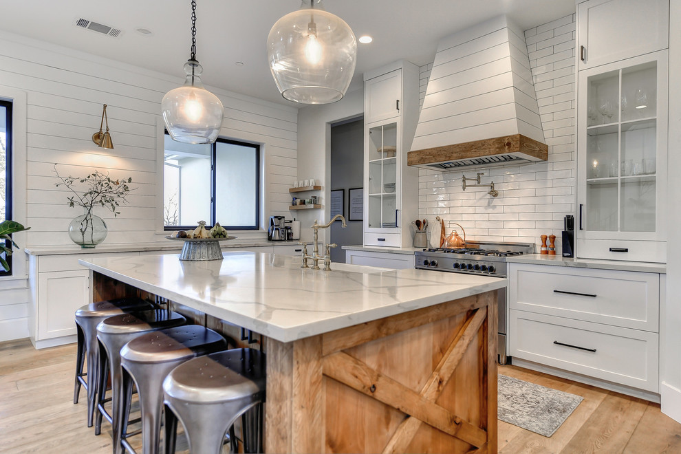 Country kitchen in Sacramento with a farmhouse sink, white cabinets, marble benchtops, white splashback, subway tile splashback, stainless steel appliances, light hardwood floors, with island, grey benchtop and shaker cabinets.