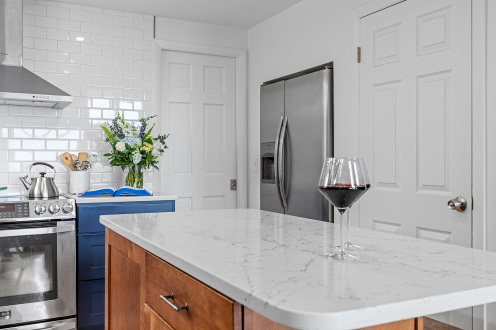 Design ideas for a transitional l-shaped kitchen in Providence with a farmhouse sink, shaker cabinets, blue cabinets, white splashback, ceramic splashback, stainless steel appliances, medium hardwood floors, with island and white benchtop.
