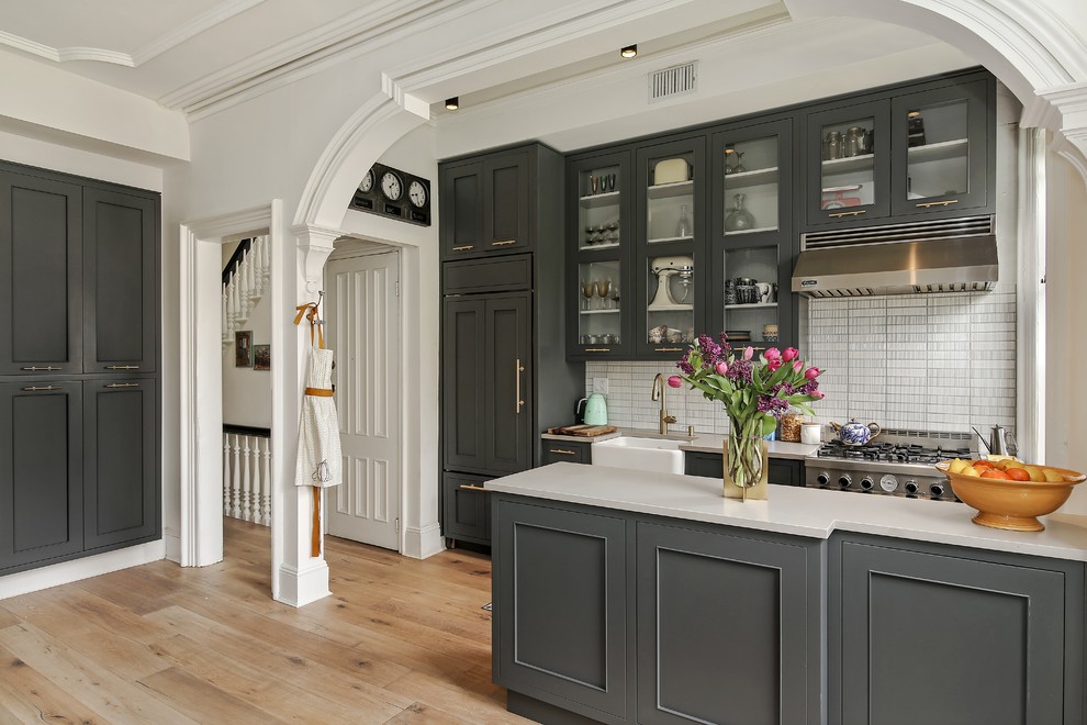 Design ideas for a mid-sized galley eat-in kitchen in Philadelphia with a farmhouse sink, recessed-panel cabinets, grey cabinets, solid surface benchtops, white splashback, ceramic splashback, stainless steel appliances, medium hardwood floors, with island and beige floor.