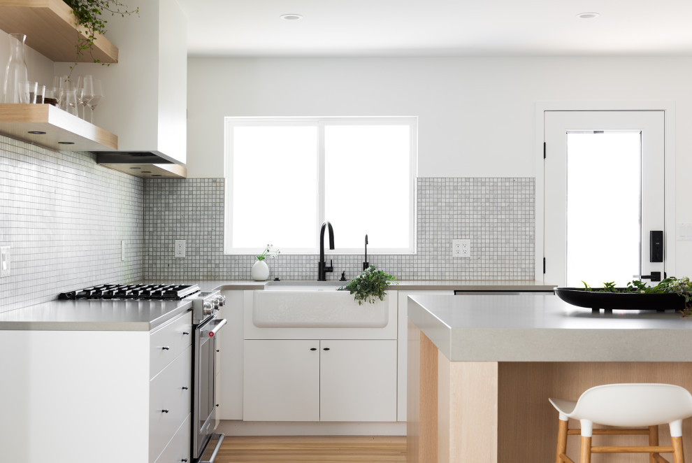 Inspiration for a small scandinavian l-shaped kitchen/diner in Salt Lake City with a belfast sink, flat-panel cabinets, light wood cabinets, engineered stone countertops, grey splashback, mosaic tiled splashback, stainless steel appliances, light hardwood flooring, an island, beige floors and grey worktops.