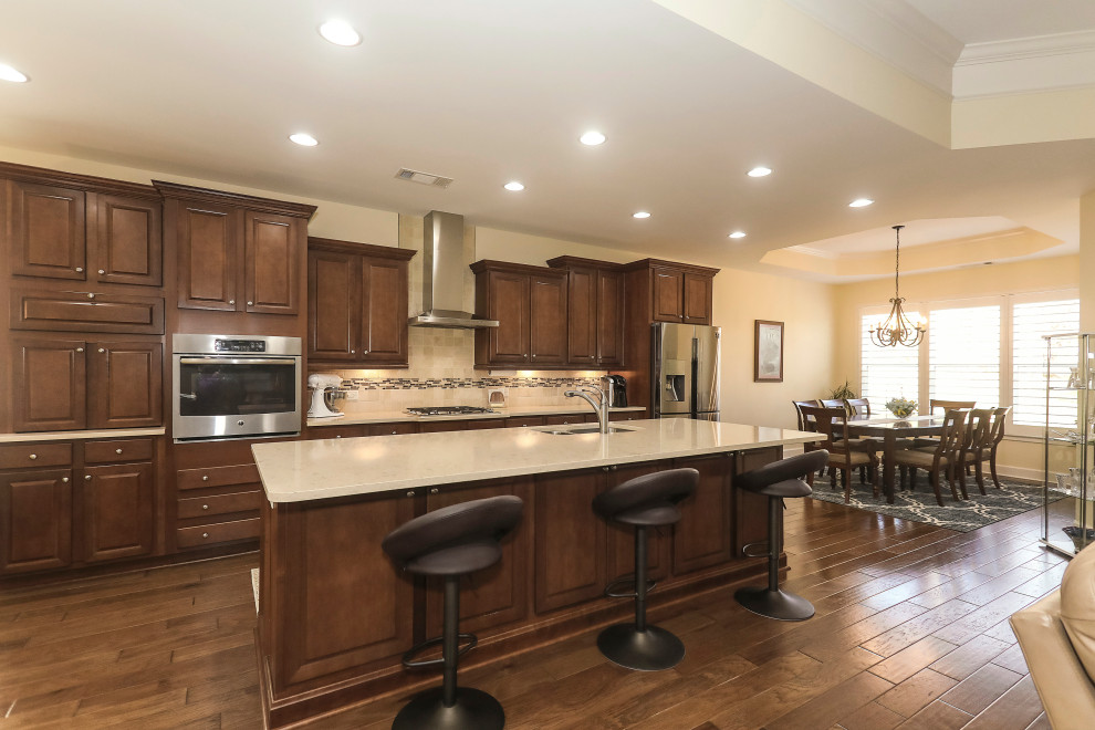Photo of a mid-sized traditional single-wall eat-in kitchen in Atlanta with a double-bowl sink, raised-panel cabinets, medium wood cabinets, quartz benchtops, beige splashback, travertine splashback, stainless steel appliances, medium hardwood floors, with island, brown floor, beige benchtop and recessed.