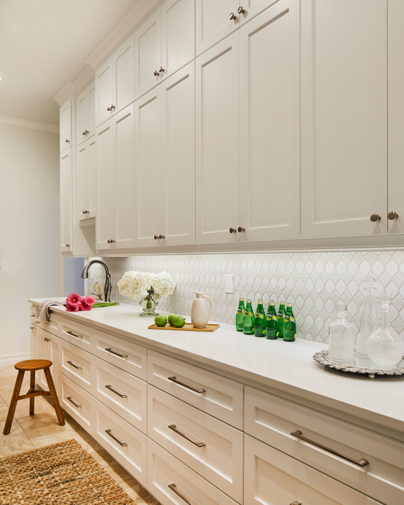 This is an example of an expansive transitional galley kitchen pantry in Other with an undermount sink, shaker cabinets, white cabinets, solid surface benchtops, white splashback, porcelain splashback, stainless steel appliances, travertine floors, beige floor and white benchtop.