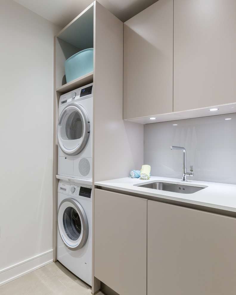 Photo of a small contemporary dedicated laundry room in Gloucestershire with an undermount sink, flat-panel cabinets, beige cabinets, solid surface benchtops, porcelain floors, a stacked washer and dryer, grey floor, white benchtop and white walls.