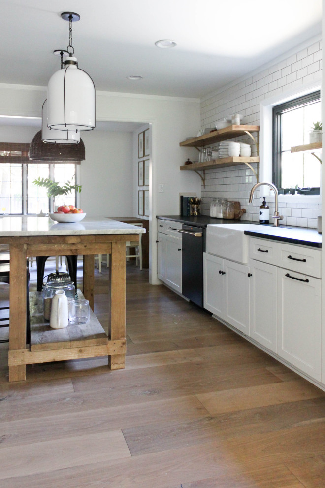 Design ideas for a mid-sized country single-wall eat-in kitchen in Indianapolis with a farmhouse sink, shaker cabinets, white cabinets, white splashback, subway tile splashback, medium hardwood floors, with island, brown floor and black benchtop.