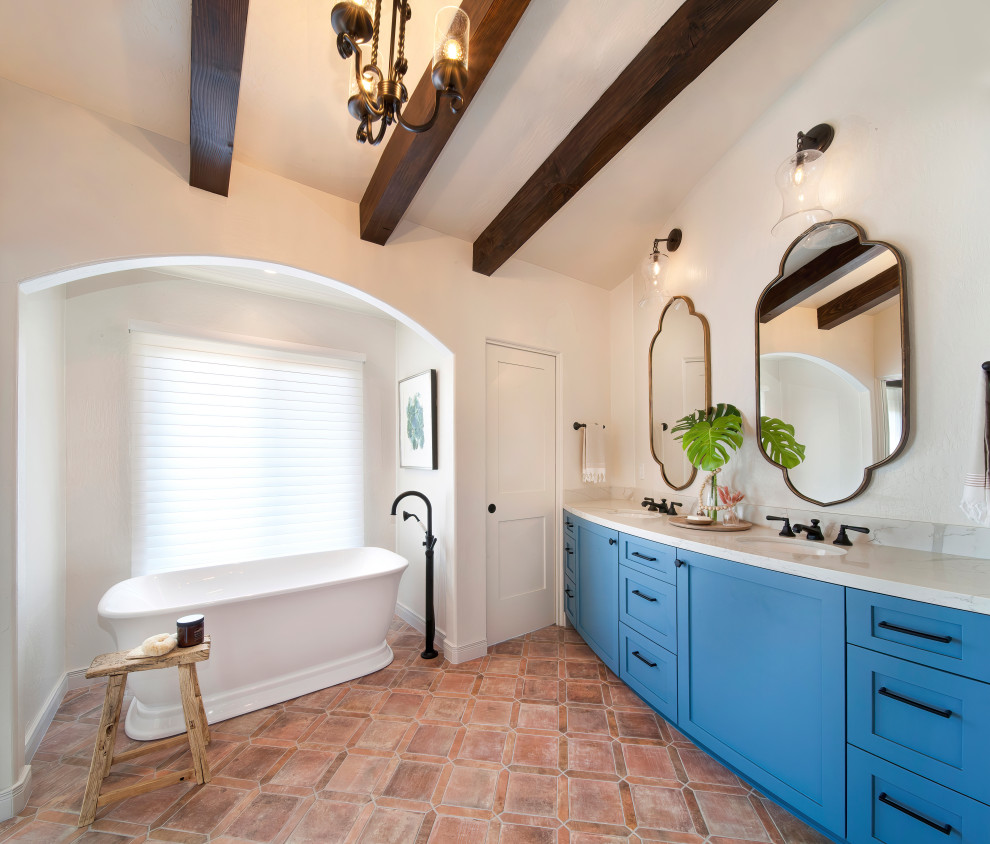 Photo of a large mediterranean ensuite bathroom in Orange County with shaker cabinets, blue cabinets, a freestanding bath, an alcove shower, a one-piece toilet, white tiles, porcelain tiles, white walls, porcelain flooring, a submerged sink, engineered stone worktops, multi-coloured floors, a hinged door, white worktops, a wall niche, double sinks, a built in vanity unit and exposed beams.