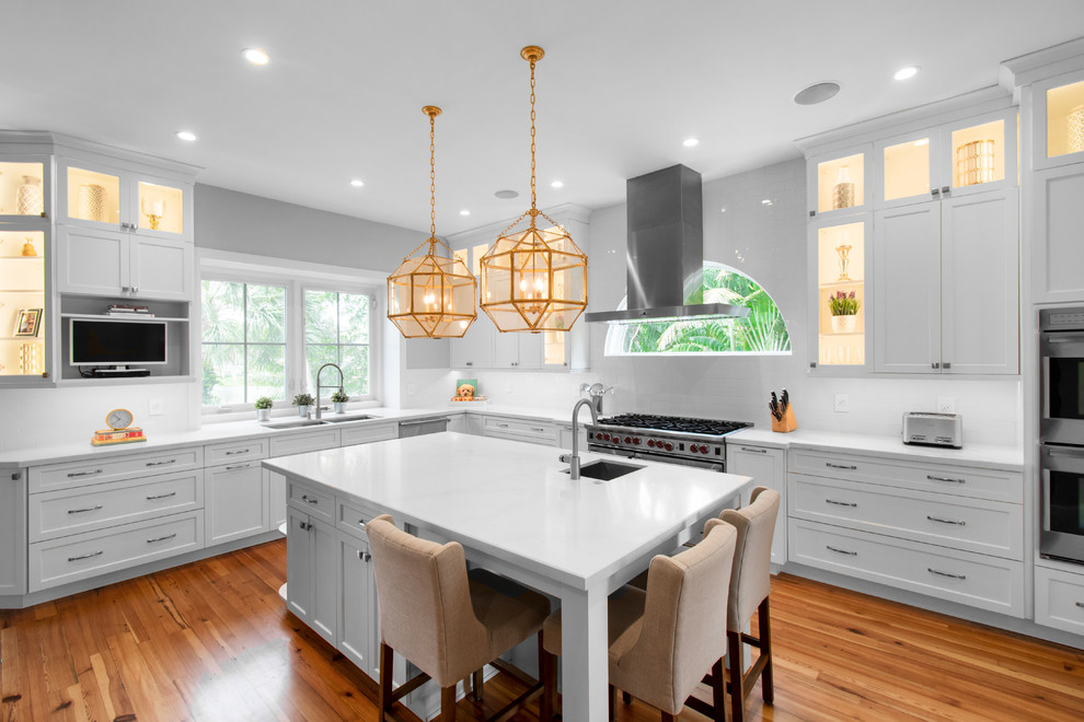 Classic l-shaped kitchen in Miami with a double-bowl sink, shaker cabinets, white cabinets, white splashback, stainless steel appliances, medium hardwood flooring, an island, brown floors and white worktops.