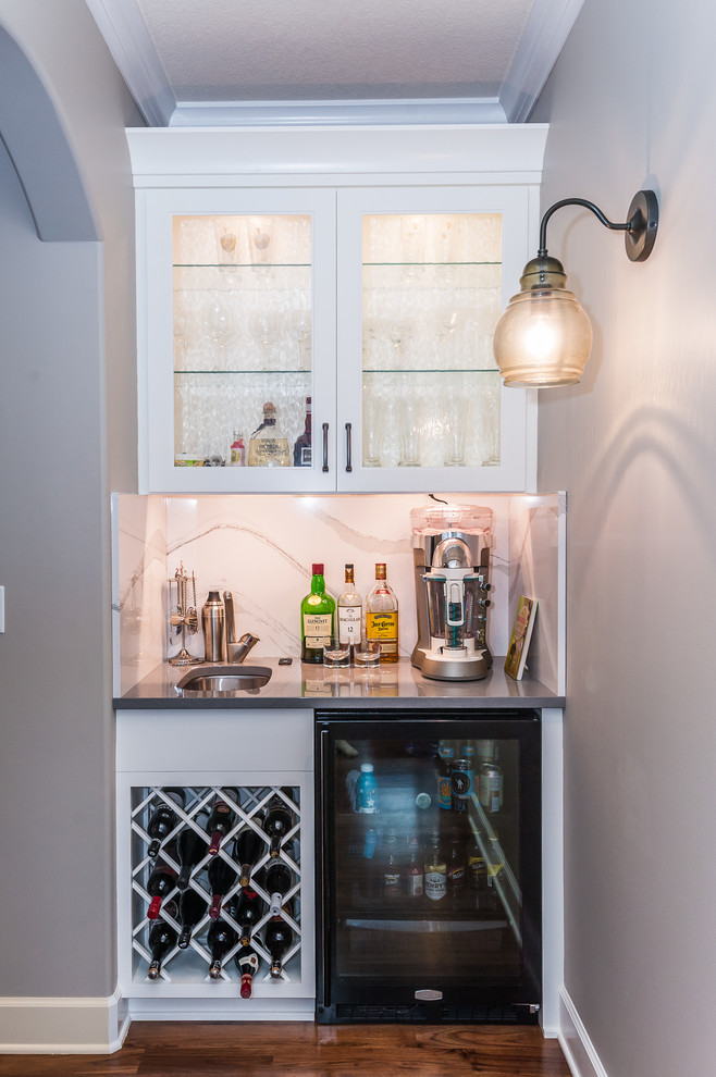 Inspiration for a small transitional single-wall wet bar in Other with an undermount sink, glass-front cabinets, white cabinets, solid surface benchtops, white splashback, stone slab splashback, dark hardwood floors and brown floor.