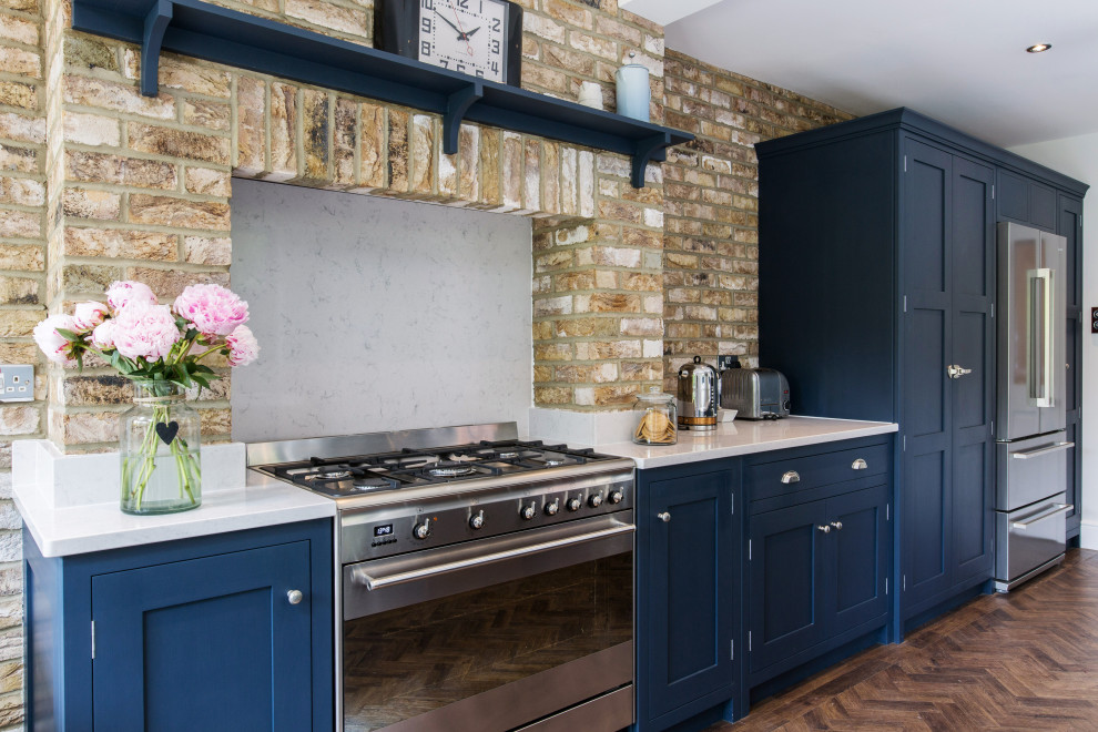 This is an example of a mid-sized industrial galley eat-in kitchen in Essex with a farmhouse sink, shaker cabinets, blue cabinets, quartzite benchtops, white splashback, brick splashback, stainless steel appliances, medium hardwood floors, no island, brown floor and white benchtop.