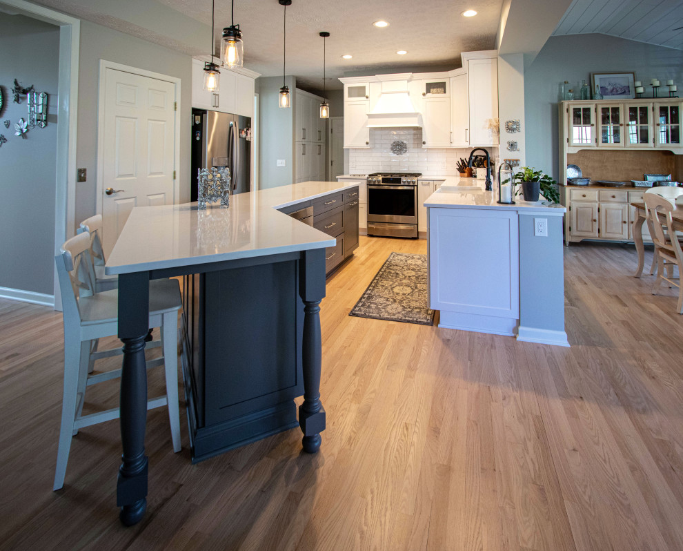 Example of a mid-sized transitional galley light wood floor and brown floor kitchen pantry design in Cleveland with a farmhouse sink, shaker cabinets, white cabinets, quartz countertops, white backsplash, ceramic backsplash, stainless steel appliances, an island and white countertops