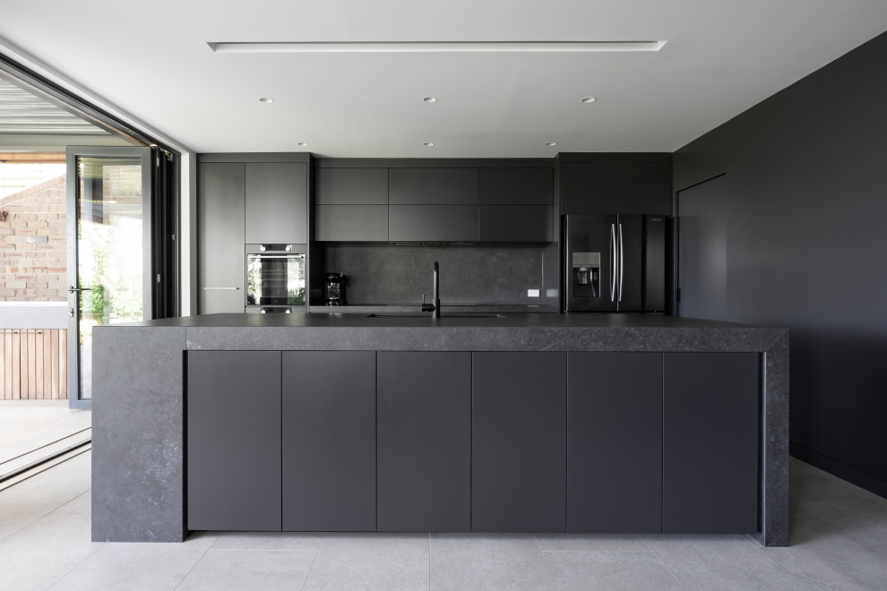 Mid-sized contemporary galley open plan kitchen in Sydney with an undermount sink, grey cabinets, solid surface benchtops, grey splashback, stone slab splashback, black appliances, cement tiles, with island, grey floor and grey benchtop.